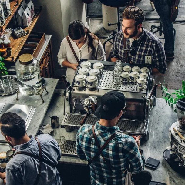 A busy coffee shop counter where two employees work on a drink order for two waiting customers