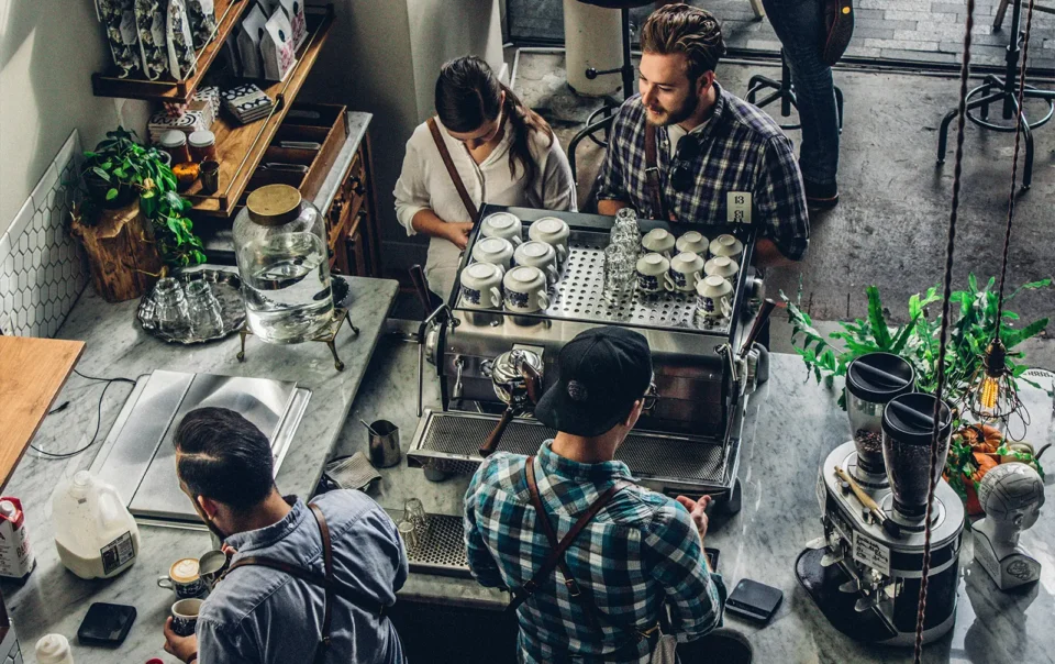 A busy coffee shop counter where two employees work on a drink order for two waiting customers