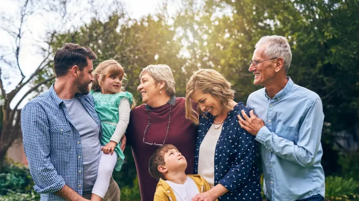 Three generations of a family pose for a photo outdoors, smiling at each other