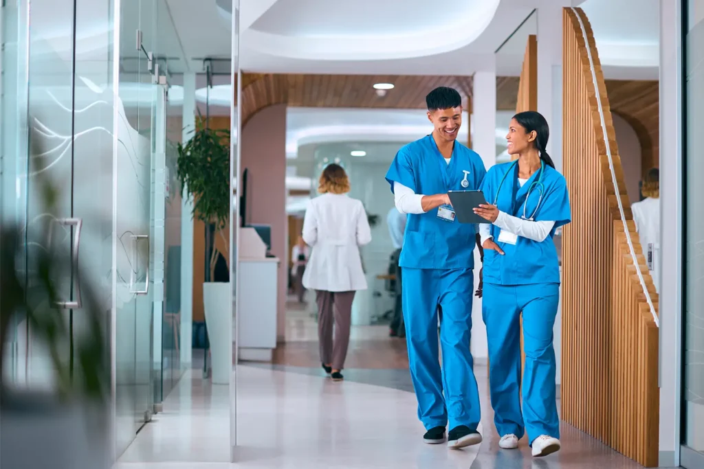 Two doctors wearing blue scrubs walking down a medical facility hallway, discussing some paperwork.