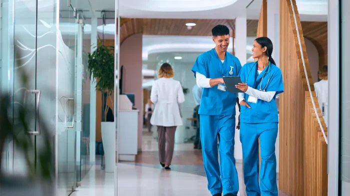 Two doctors wearing blue scrubs walking down a medical facility hallway, discussing some paperwork.