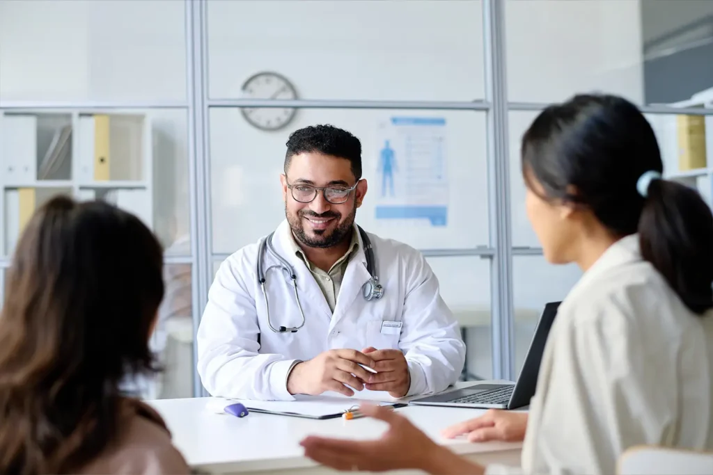 A male doctor faces the camera, smiling at two patients seated across from him.