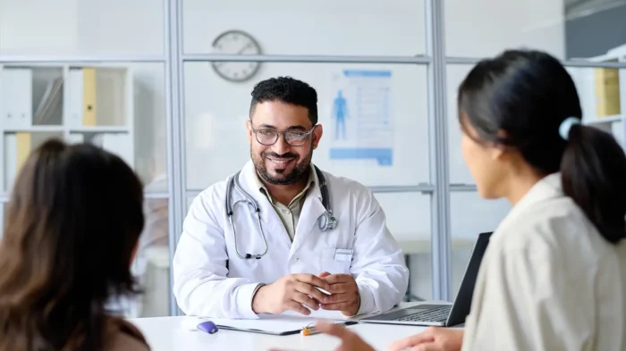 A male doctor faces the camera, smiling at two patients seated across from him.
