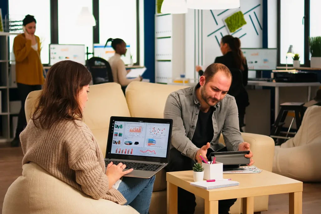 In a startup office, a woman sits with her back to us, working on a laptop, while a man works on his tablet nearby.