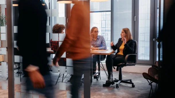 Three workers have a discussion in a private board room as two blurry employees walk by