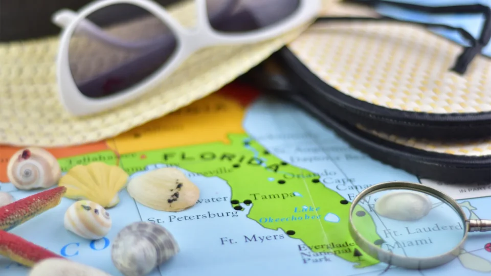 A colorful map of the United States, focused on Florida, sits on a table. Vacation items like sea shells, white sunglasses, and sandals rest on top of it.