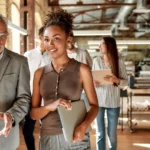 An older businessman in glasses gestures as he talks to a younger business woman walking beside him holding a laptop. Three other young professionals trail behind them. They are in an open floor plan office with exposed brick and metal.