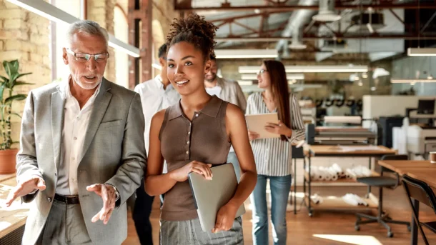 An older businessman in glasses gestures as he talks to a younger business woman walking beside him holding a laptop. Three other young professionals trail behind them. They are in an open floor plan office with exposed brick and metal.