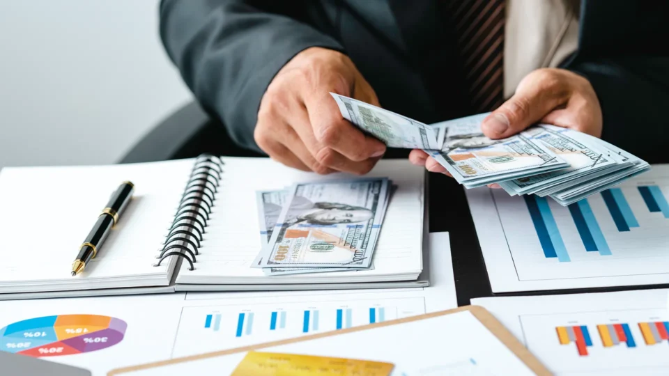 Close-up of a man's hands counting $100 bills. On the desk below, business charts and spreadsheets are arrayed on a desk.