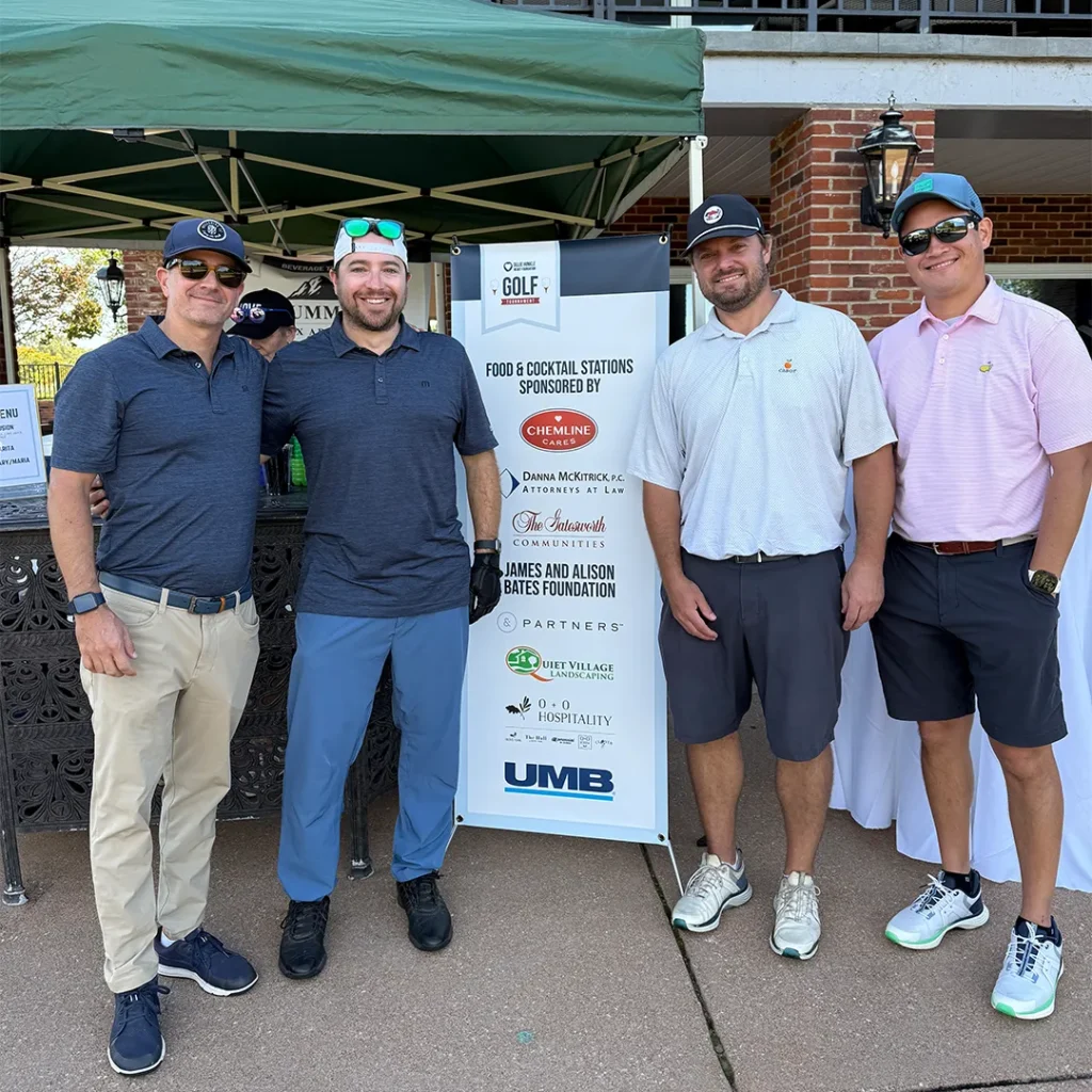 Four men in golf attire stand near a standing sign denoting the sponsors of the Ollie Hinkle Heart Foundation's charity golf tournament